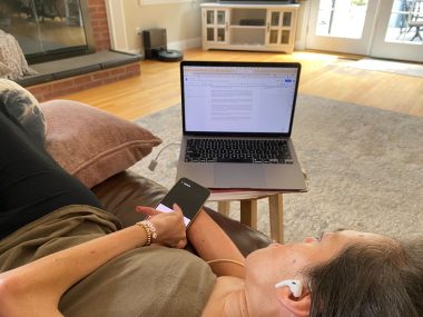 body position | SMA News Today | Alyssa lies on the couch to write her column. She's wearing AirPods and holding her phone, and her computer is open on a stool next to her.