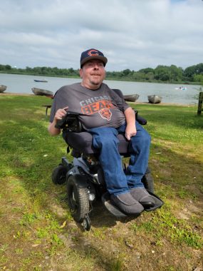 attitude and motivation | SMA News Today | Steve poses in his power wheelchair in front of a river. He is wearing a Chicago Bears T-shirt and hat
