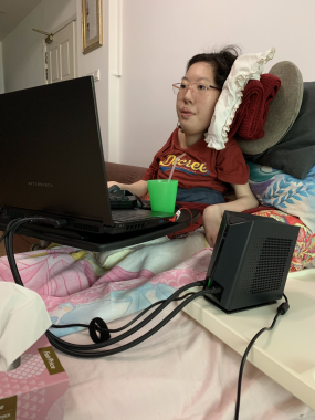 grief and trauma | SMA News Today | a photo of Sherry seated in front of her laptop and laptop's cooler, playing a game she was commissioned to review. Six pillows lie behind her, keeping her upright.