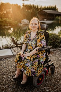 SMA News Today | Stéphanie Houvet wears a floral patterned dress and sits in her wheelchair near a small pond at sunset.