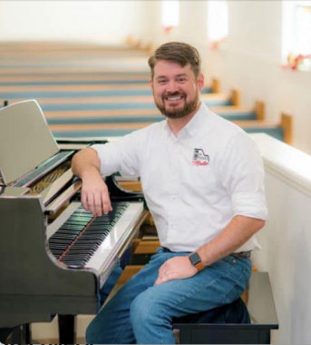 The picture shows a young man at the side of a piano with church pews behind him and white walls. He wears a white shirt with an insignia and blue jeans. He has brown hair and a beard and mustache.
