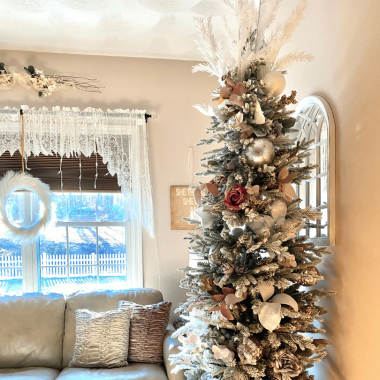 A white and green beautifully decorated Christmas tree sits in a stylish bedroom with beige and white color tones. A white wreath hangs in the window below white curtains and white Christmas lights.