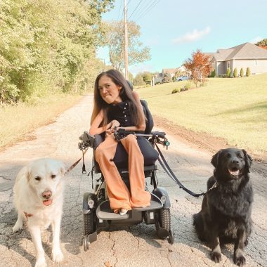 A woman strolls with her two dogs down a peaceful country or suburban lane, with trees and scattered houses in the background. The woman is in the center, in a power wheelchair, and to her right is a large golden retriever on a leash, and to her left, a black golden retriever, also on a leash. 