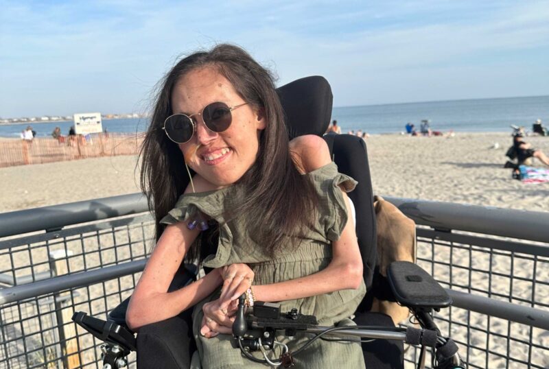 A woman with long brown hair poses at the beach in a green romper and dark sunglasses. She's seated in her power wheelchair in front of a metal railing, with a stretch of sand and the ocean visible behind her.