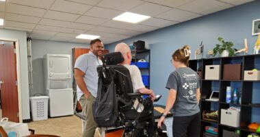 A man raises his wheelchair as his physical therapist and another physical therapist watch.