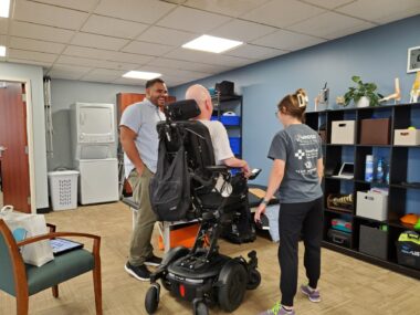 A man raises his wheelchair as his physical therapist and another physical therapist watch.