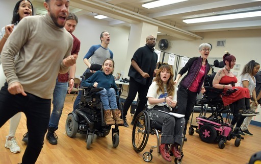 Multiple people, some using wheelchairs, sing together in a large open room.