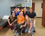 A group of people surround one man in a wheelchair, all are wearing birthday hats and smiling.