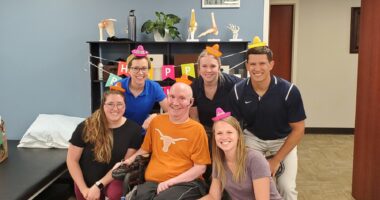 A group of people surround one man in a wheelchair, all are wearing birthday hats and smiling.