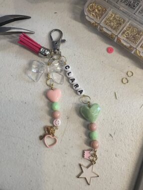 A keychain being made sits on a workbench, with a box of jewelry items to the upper right, some type of tool (like a pair of scissors) to the left, and then a keychain with two strands. Heart-shaped beads and round beads in pink and lime green accent the strands of the keychain, and toward the top letters spell out the name "Clara." 