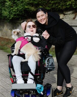 A woman leans over a teenage girl in a wheelchair who has a dog perched on her lap.