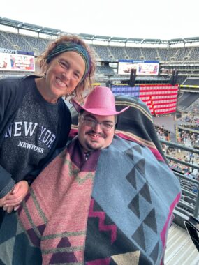 A mother and her adult son smile broadly from their seats at a stadium ahead of a Beyoncé concert. The mom is wearing a New York sweater while the man sports a colorful, patterned poncho and a pink cowboy hat.