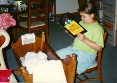 A young girl, about 7 or 8 years old, sits in a wooden chair reading a children's book. Her baby brother lies in a wooden bassinet in front of her.