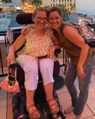 Two women smile and lean together for a side hug. One of the women is seated in a power wheelchair, and the other is standing beside her, slightly bent over to wrap her arm around her friend's shoulders.