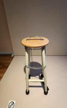A horizontal close-up photo from a phone showing a common wooden stool with a handle affixed to the top and a metal chain wrapped around it. It is sitting in a gallery display. 