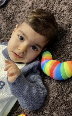 A young boy lies on a rug with a colorful toy by his head. He looks up at the camera and smiles. 